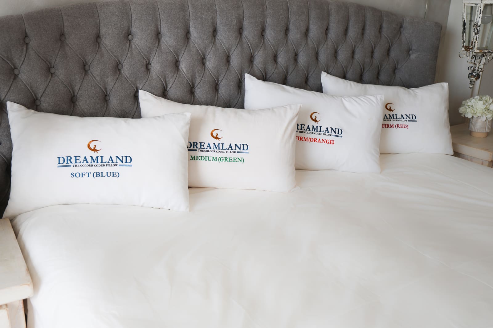 Four Dreamland pillows lined up on a bed against a grey tufted headboard, with blue, green, orange and red embroidered labels