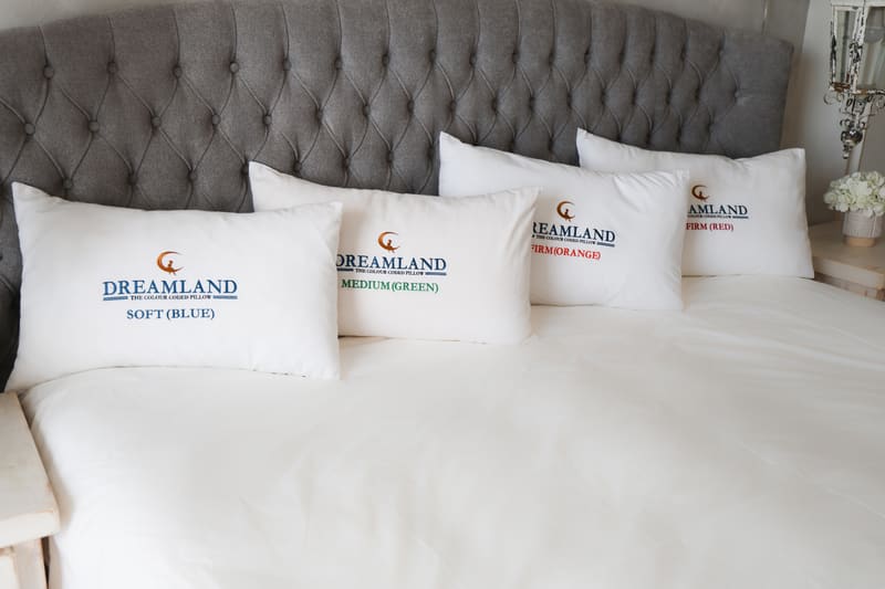 Four Dreamland pillows lined up on a bed against a grey tufted headboard, with blue, green, orange and red embroidered labels
